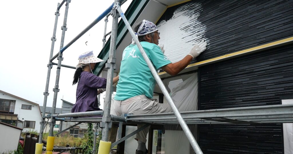 足場に二人並んで腰かけ、正面の壁にMORUMORUを塗っている様子